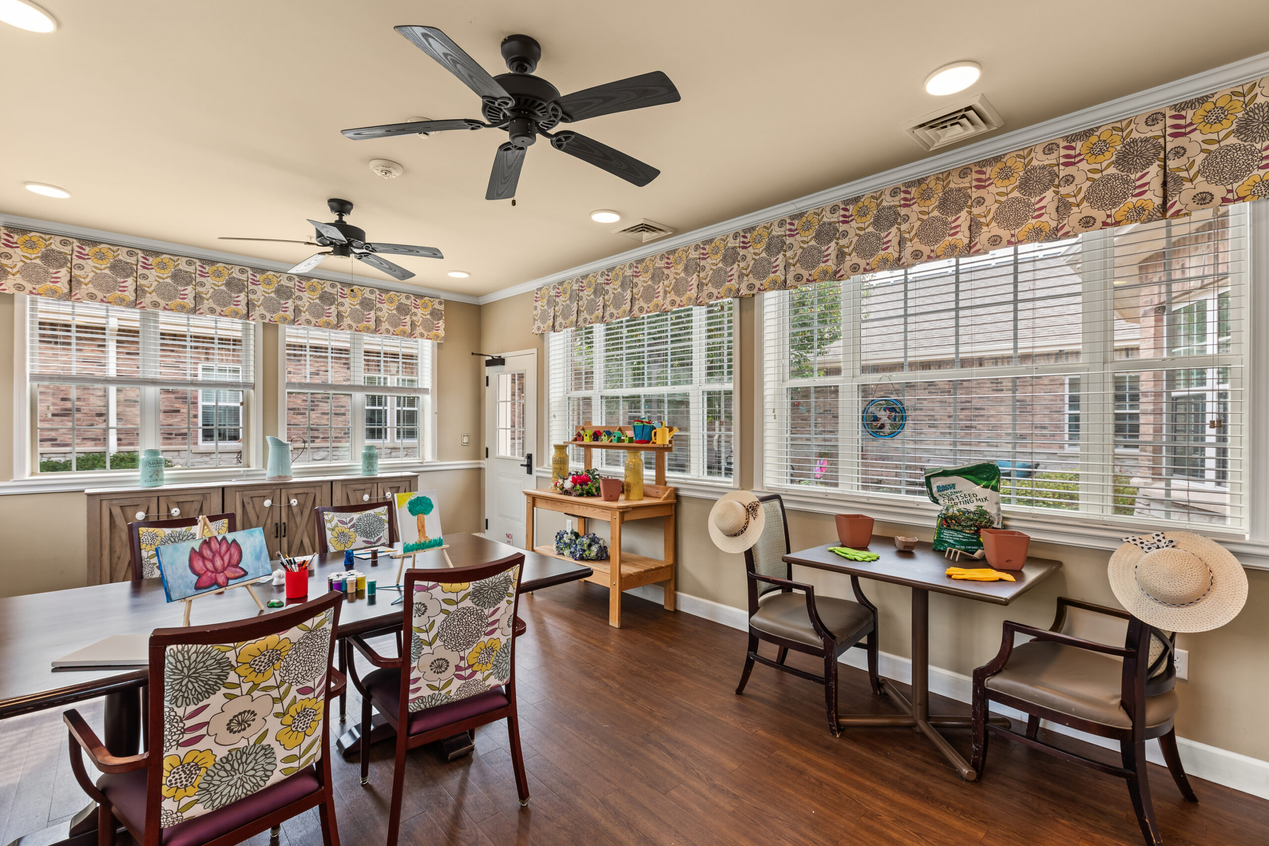 A bright activity or art room with large windows, light yellow walls, dark wood floors, ceiling fans, and a long table set up for painting and crafts.