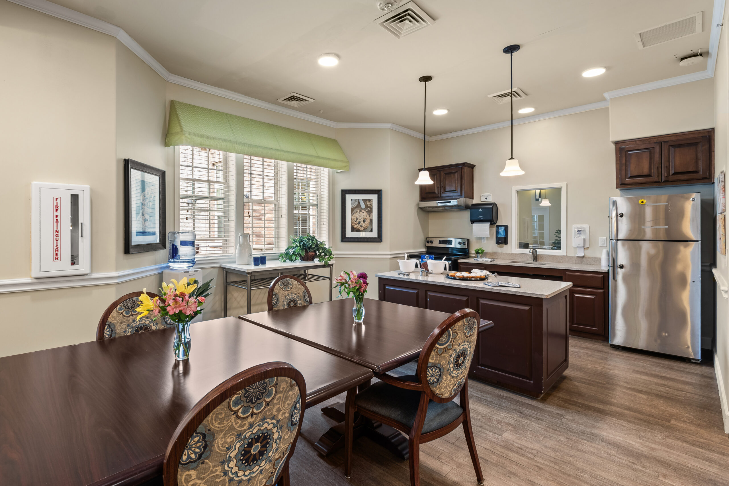 A cozy, well-lit bistro area with a small kitchen featuring dark cabinets and stainless steel appliances, a center island, and a dining table set with flowers.