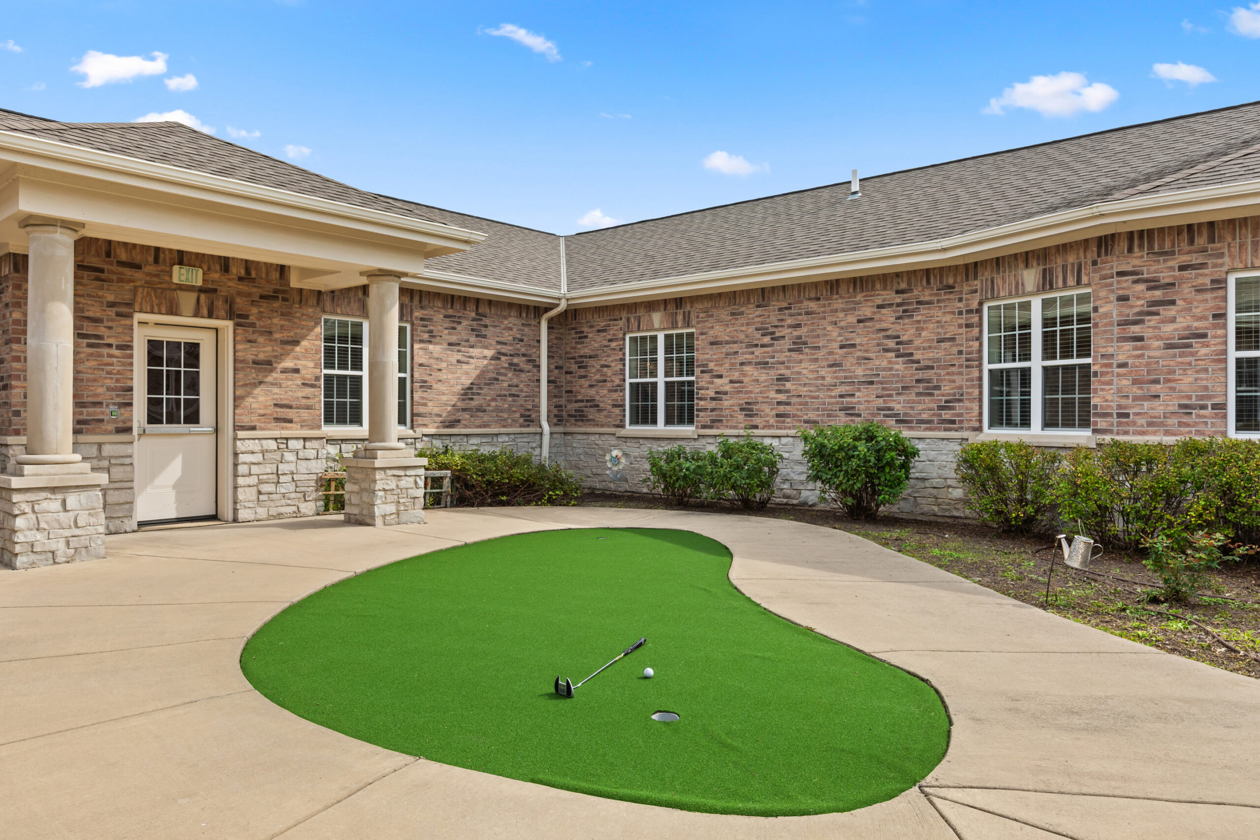 A small, outdoor putting green shaped like a kidney bean, set on a paved patio surrounded by a brick building. A golf ball and putter are visible on the turf.