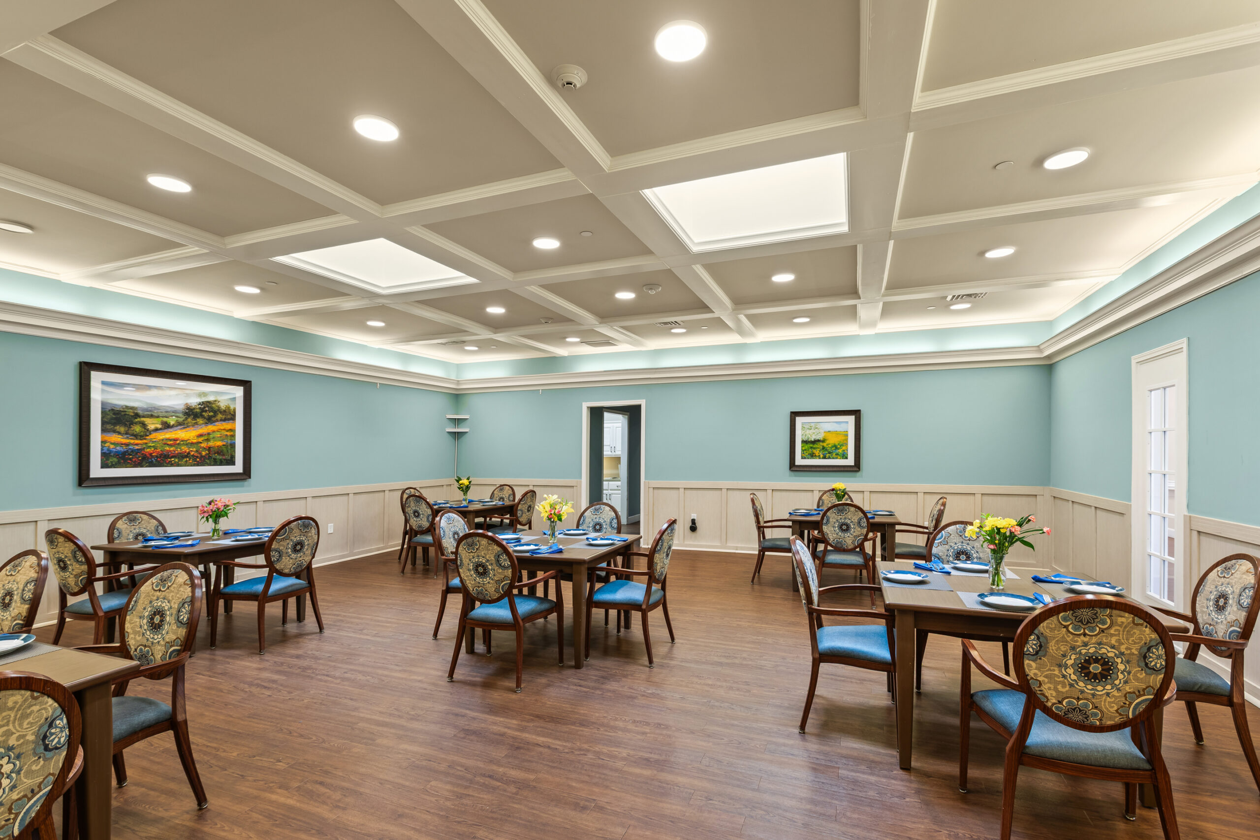 A wide view of a spacious, well-lit dining room with light blue walls, white trim, dark wood tables set for dining, and framed landscape artwork on the walls.