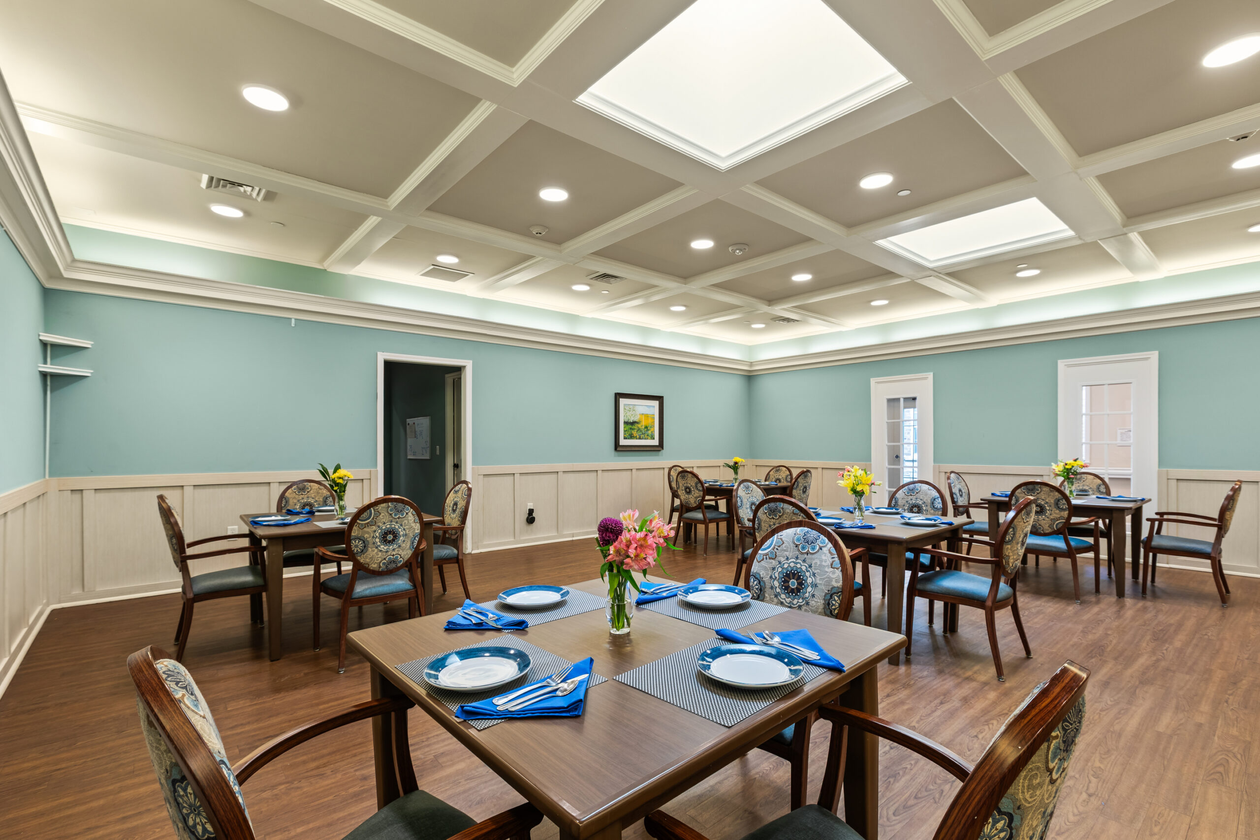 A formal dining room in a senior living community with light blue walls, white wainscoting, a coffered ceiling, and individual tables set with blue napkins and floral centerpieces.