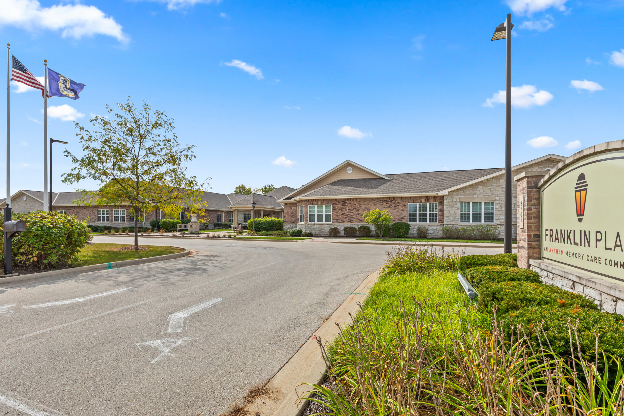 An exterior view of a single-story brick and stone assisted living or memory care community building, with a road leading to the entrance and a large sign partially visible on the right. American and state flags are flying.