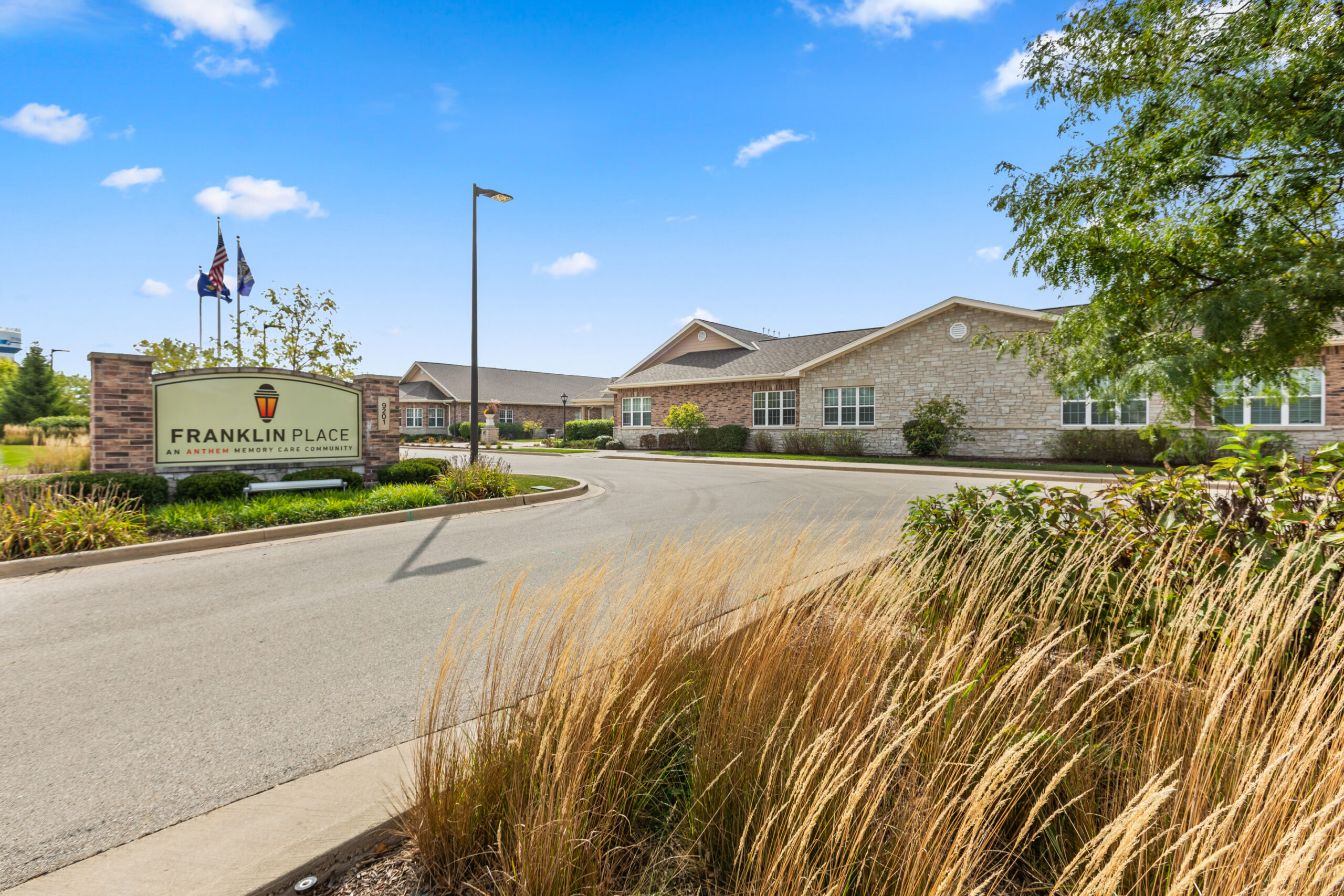 An exterior view of the "Franklin Place" senior living community sign and entrance, showing the long, low brick and stone building under a bright blue sky.