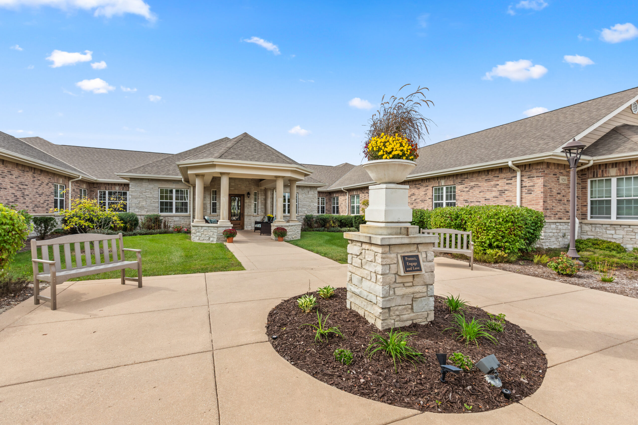 The main walkway and front entrance of a senior living community, featuring a decorative stone planter with yellow flowers, wooden benches, and a wide paved path leading to the entrance.