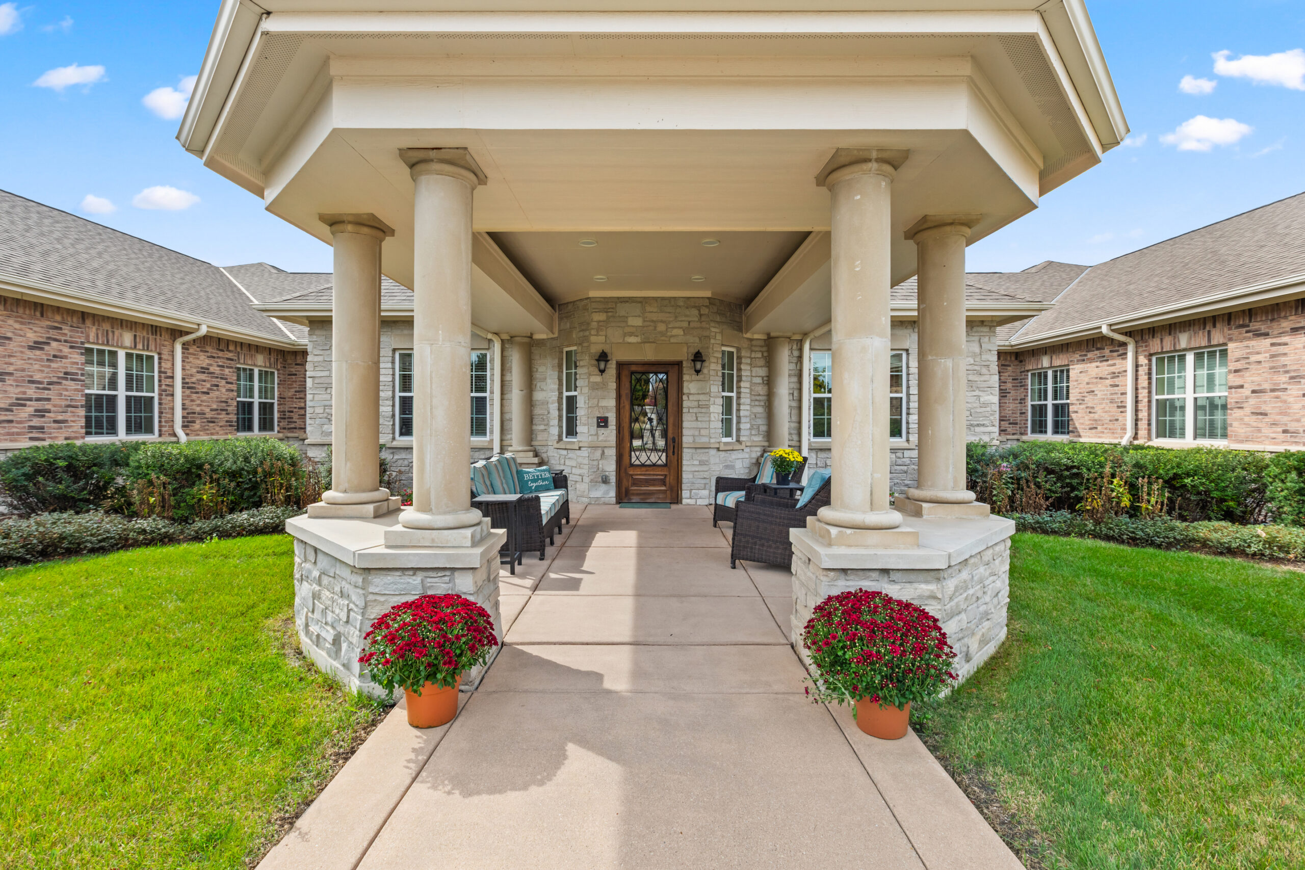 The main covered entrance of a senior living facility, featuring a portico with large stone columns, two wicker seating areas, and planters with bright red flowers.