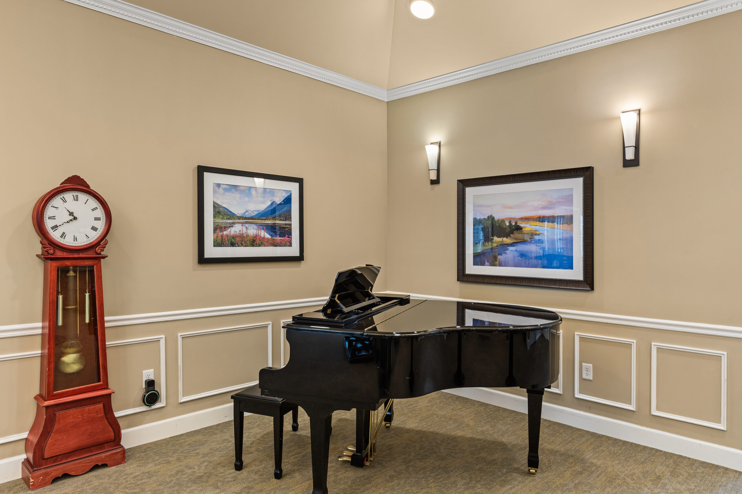 A formal music corner with a glossy black grand piano and bench, a large red grandfather clock, and two framed landscape pictures on the beige walls.