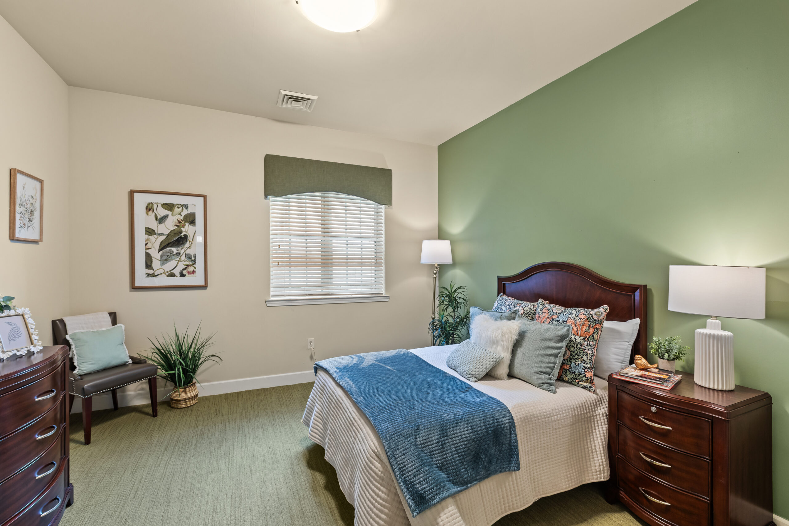 A model bedroom with light beige walls, one olive green accent wall, green patterned carpet, dark wood furniture, and a full-sized bed with white and blue bedding.