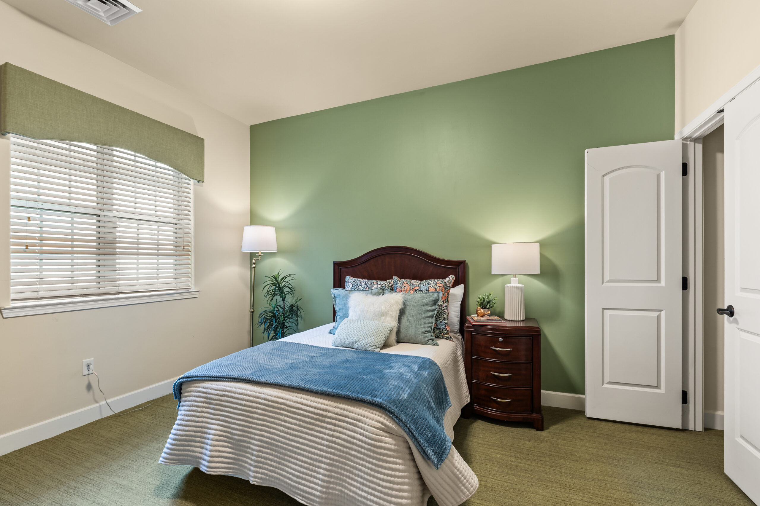 A model unit bedroom with an olive green accent wall behind a dark wood bed, a large window with white blinds, and a blue throw blanket over white bedding.