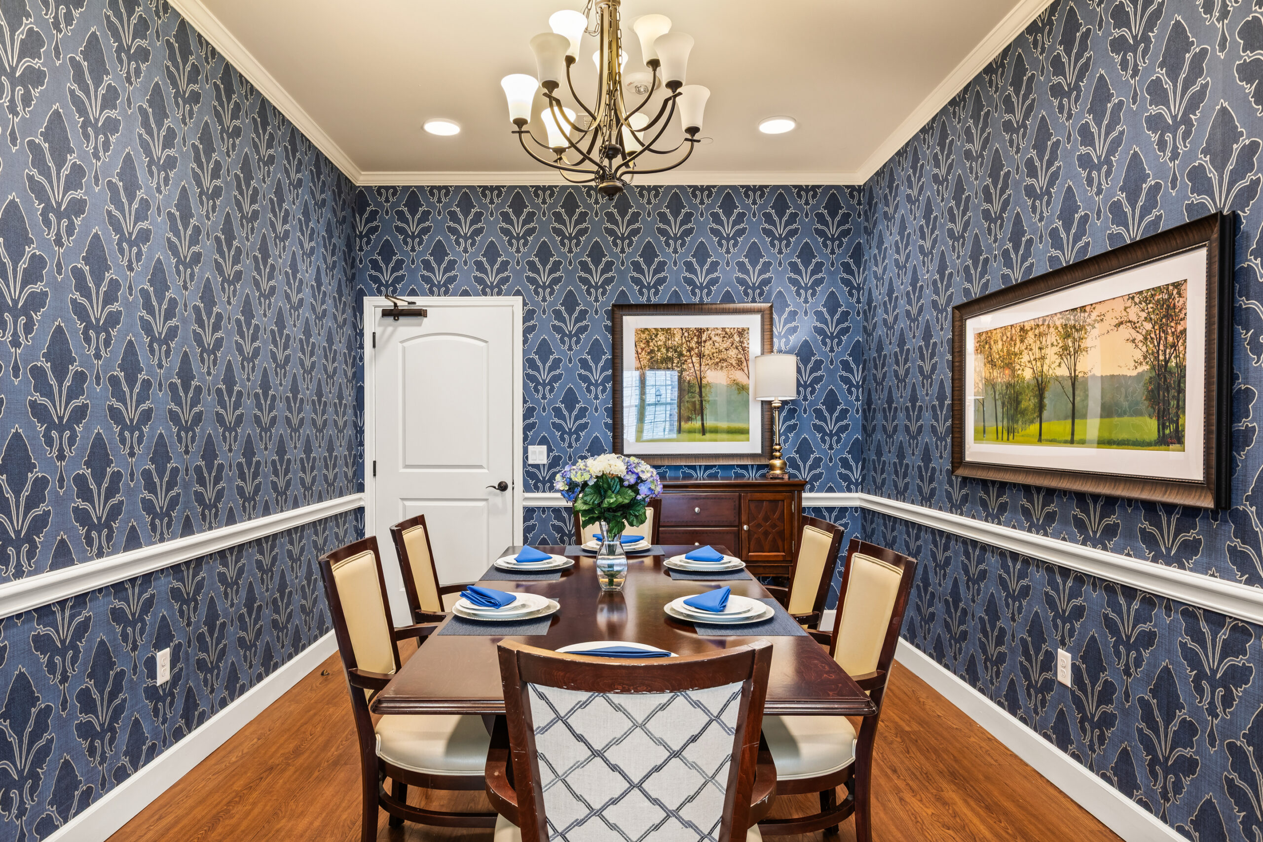 A private dining room with bold navy blue and white damask wallpaper, white wainscoting, a dark wood table set for six, and two landscape paintings.