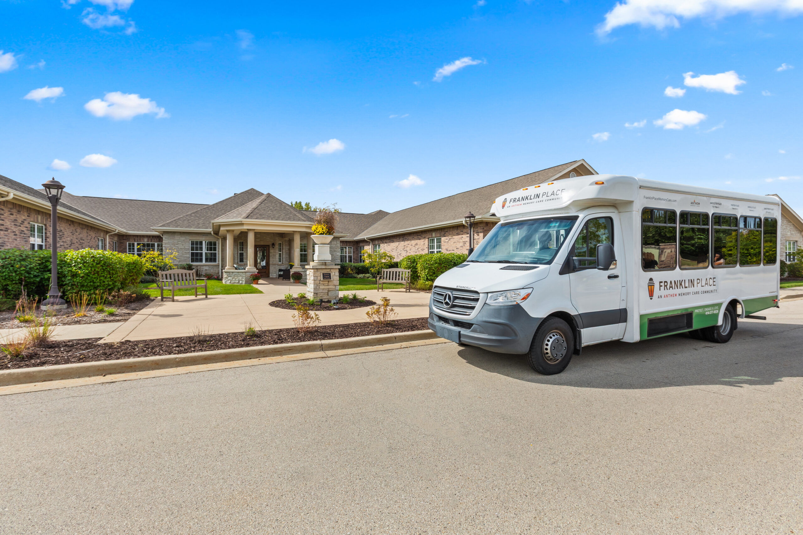A white passenger van with "Franklin Place" branding parked in front of the senior living community building, under a bright blue, partially cloudy sky.
