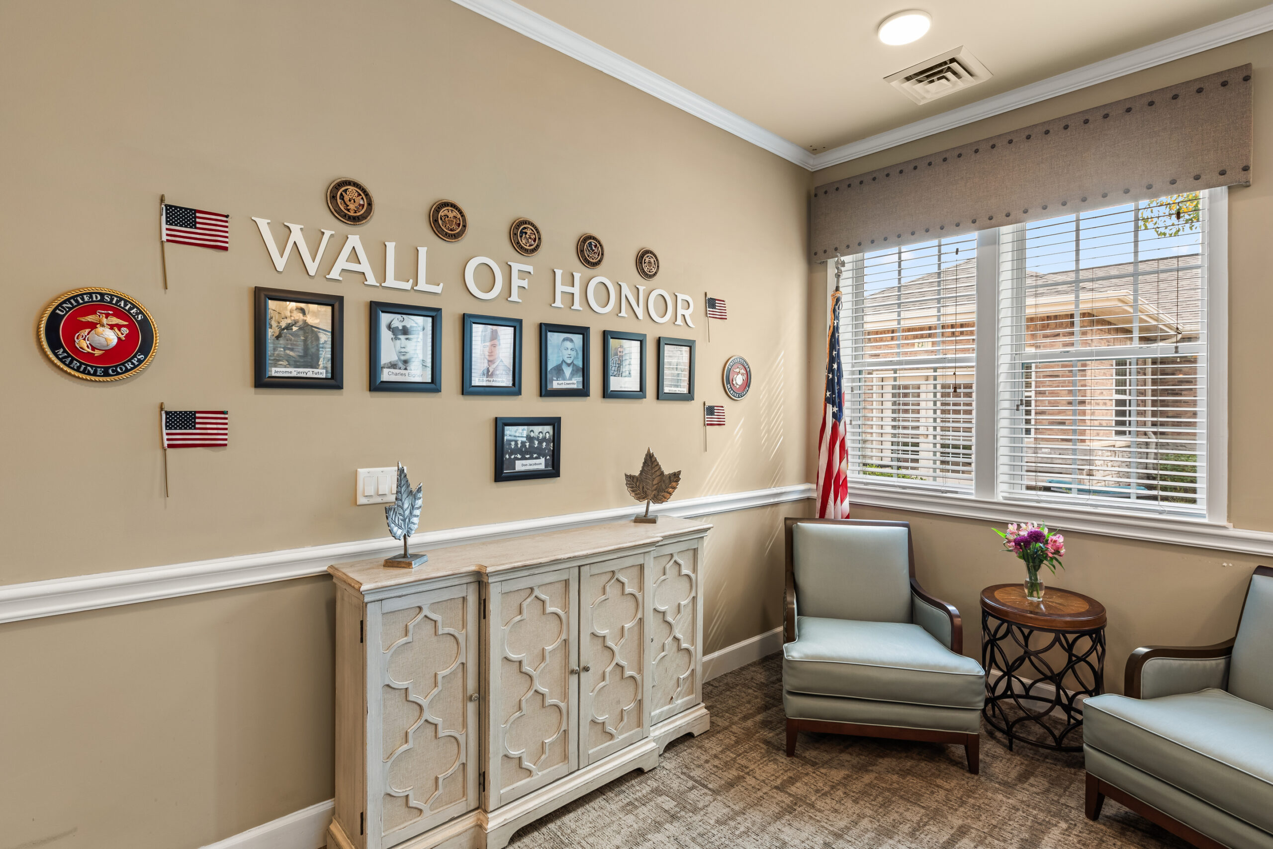 A "Wall of Honor" in a community room featuring framed photos of veterans, military emblems, small American flags, and a decorative white cabinet below.